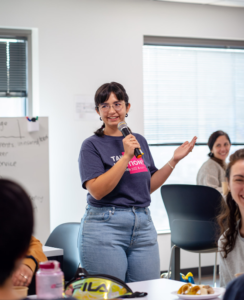 L&T Consultant Gabriela emcees the event, speaking on a mic in front of the group of hackers in a blue shirt and jeans.