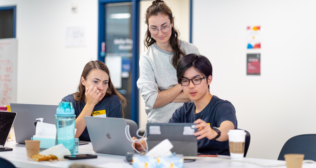 Two hackathon participants sit at laptops working on something. over their shoulder, an event coordinator is looking at their screen to see what they're working on.